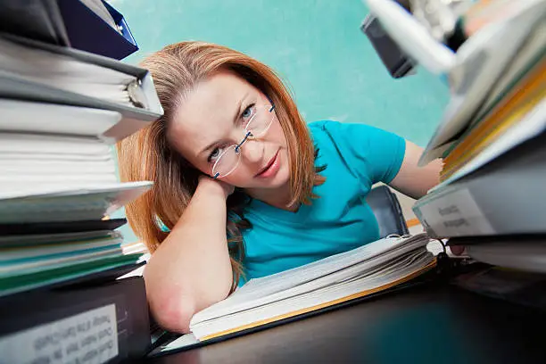 Pretty mid adult Caucasian teacher is stressed as she grades papers in her classroom. Piles of books and note books are on her desk. A chalkboard is in the background.