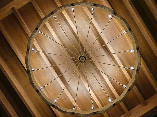 A circular chandelier is photographed from below and is viewed against a timber beamed ceiling which it is illuminating.