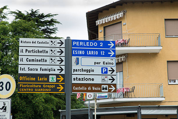 Varenna, Italy. Sign indicating the road to Esino Lario Varenna, province of Lecco, Italy - May 27, 2016: road signs in Varenna: on the right, in blue, are indicates the suburban roads to go to two villages near Varenna: Perledo (2 km - 1,2 miles) and Esino Lario (12 km - 7,5 miles), we are in fact at the fork road that leads to the beginning of the climb for the small tourist village of Esino Lario located to 913 meters above sea level. In white, are indicates a car park and the station; in brown I have a tourist sign indicating the presence of a castle.To the left, in black, are indicated two industrial activities, a workshop for car and a floriculture; the remaining white signs indicate the presence of taverns or restaurants. wikipedia stock pictures, royalty-free photos & images
