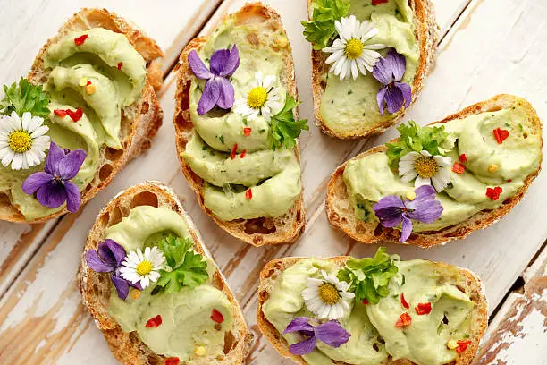 Delicious canapes with avocado paste and edible flowers on wooden background
