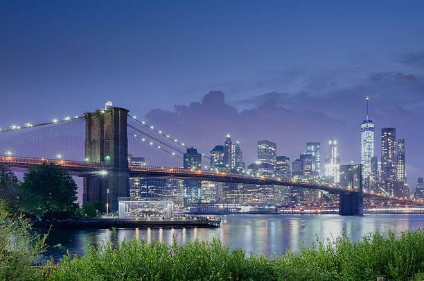 A long exposure photo of the world famous New York City Skyline and the Brooklyn Bridge that spans the East River. In the background you see the Manhattan skyline with the financial district, Wall Street and the World Trade Center Freedom Tower in view. In the foreground we have the Brooklyn Bridge Park with people blurred out by movement.