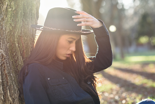 Portrait of thoughtful woman sitting alone outdoors wearing hat. Nice backlit with sunlight