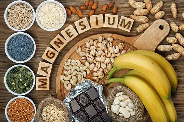 Products containing magnesium: bananas, pumpkin seeds, blue poppy seed, cashew nuts, beans, almonds, sunflower seeds, oatmeal, buckwheat, peanuts, pistachios, dark chocolate and sesame seeds on wooden table