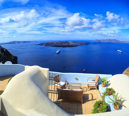 Panoramic view of Santorini volcanic islands from terrace with garden furniture and plants in pots. Cruise ship on water. Clouds in sky.