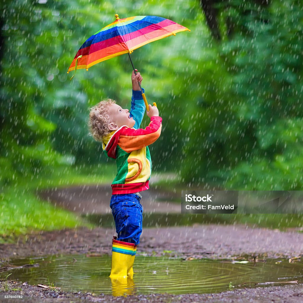 Little child playing in the rain Little boy playing in rainy summer park. Child with colorful rainbow umbrella, waterproof coat and boots jumping in puddle and mud in the rain. Kid walking in autumn shower. Outdoor fun by any weather Rain Stock Photo