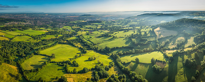 Gentle mist disappearing in the green folds an idyllic rural environment overlooking a vibrant patchwork quilt of meadow and pasture, country town and hedgerows from high above in this picturesque aerial panorama of the iconic Cotswolds landscape, Gloucestershire, UK. ProPhoto RGB profile for maximum color fidelity and gamut.