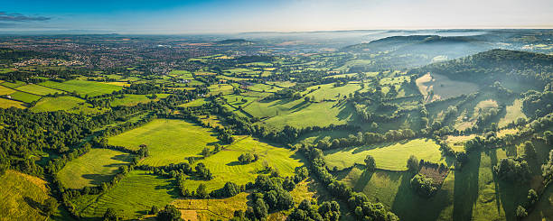 Aerial panorama over green fields misty hills and country town Gentle mist disappearing in the green folds an idyllic rural environment overlooking a vibrant patchwork quilt of meadow and pasture, country town and hedgerows from high above in this picturesque aerial panorama of the iconic Cotswolds landscape, Gloucestershire, UK. ProPhoto RGB profile for maximum color fidelity and gamut. gloucestershire stock pictures, royalty-free photos & images