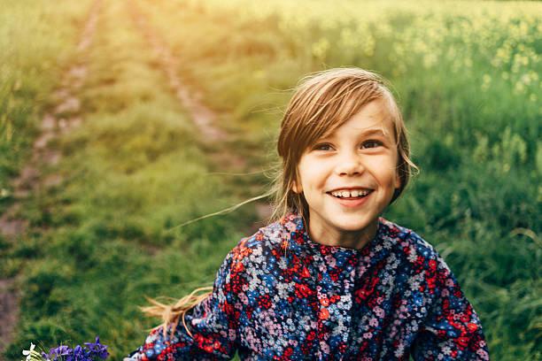 11-year-old girl holds out a blueberry in her hand, smiling widely at the camera. She wears a white summer dress while standing in a green field near a lake. The background shows blueberry bushes and the golden light of late afternoon sun. Her 19-year-old autistic brother is nearby as they spend time together gathering berries in this natural setting.