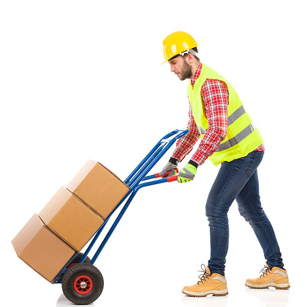 Walking delivery man Serious man in yellow hardhat and lime reflective vest pushing a delivery cart. Full length studio shot isolated on white. mover delivery person box hand truck stock pictures, royalty-free photos & images