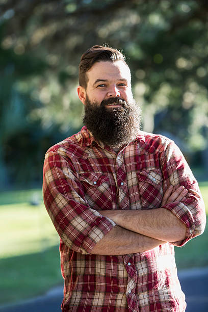 Man with beard wearing plaid shirt Portrait of a mid adult man with a full beard, wearing a lumberjack shirt, standing outdoors with arms folded. man beard plaid shirt stock pictures, royalty-free photos & images