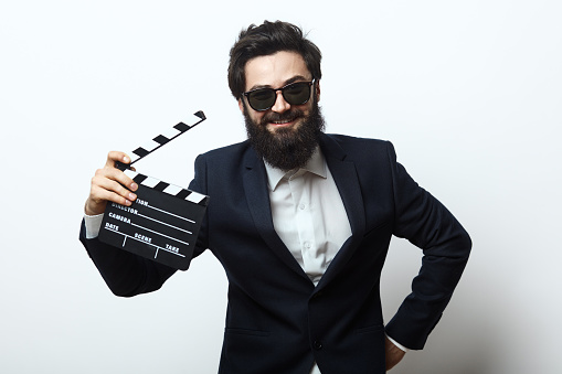 Smiling man with beard and sunglasses wearing an elegant black suit holding holding film clapper over white background. Film director concept.