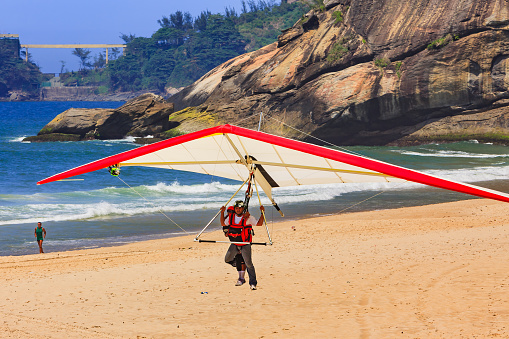 Rio de Janeiro, Brazil - November 07, 2010: A professional Hang Glider Pilot, brings a tourist in for landing on Sao Conrado Beach in Rio, after jumping off a nearby mountain to give the tourist a Hang Gliding experience and an arial view of the city.  To the starboard side of the wing is strapped a camera to give the tourist a short video of the experience.  In the background is the Atlantic Ocean.  A jogger is seen jogging on the beach quite unconcerned by the Hang Glider.  Photo shot in the afternoon sunlight; horizontal format.