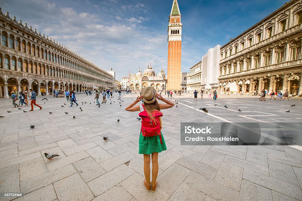 Traveling in Venice Young female traveler with hat and backpack standing on San Marco square with tower and basilica on the background in Venice. Back view with copy space Venice - Italy Stock Photo