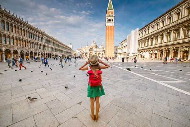Traveling in Venice Young female traveler with hat and backpack standing on San Marco square with tower and basilica on the background in Venice. Back view with copy space basilica photos stock pictures, royalty-free photos & images