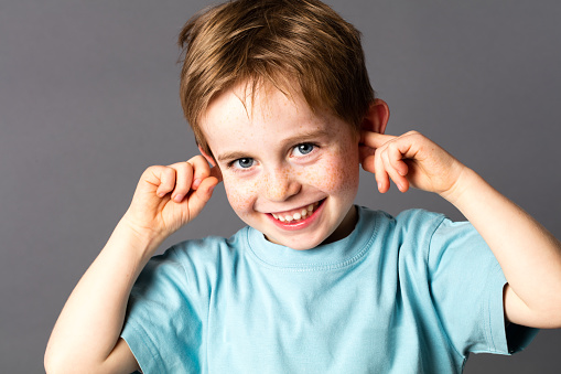 smiling cheeky young boy with blue eyes and freckles teasing, covering his closed ears, ignoring his parent scolding with attitude, grey background