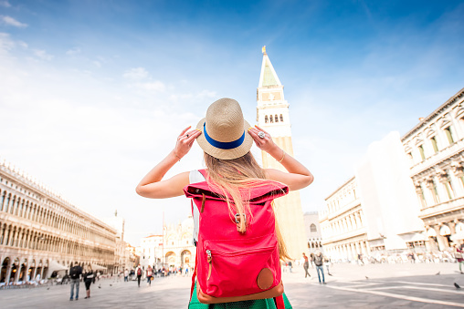 Young female traveler with hat and backpack standing on San Marco square with tower and basilica on the background in Venice. Back view