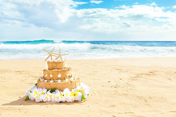 Wedding cake with starfish cake topper for a destination wedding on a tropical beach paradise. A sand castle wedding cake decorated with seashells and starfish cake topper, surrounded by a flower lei on a tropical paradise beach. Concept photo for destination wedding, and honeymoon vacation. Photographed on location in horizontal format with copy space.