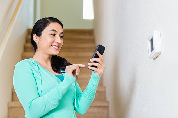 Hispanic woman adjusts thermostat with smart phone Pretty mid adult Hispanic woman uses smart phone to control temperature of home. She is standing in front of the thermostat on the wall in front of the stairs that lead to the second floor of her home. She smiles confidently as she uses the technology. She has brown hair pulled back into a pony tail and is wearing a light green hoodie. smart thermostat stock pictures, royalty-free photos & images