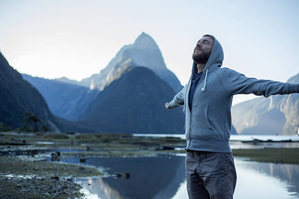 Milford Sound: Young male arms outstretched Cheering young male stands arms outstretched in front of the Mitre peak in Milford sound, New Zealand. Mitre peak reflections on the water. mitre peak stock pictures, royalty-free photos & images