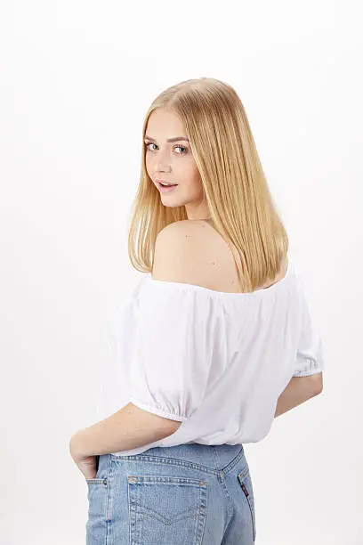 An attractive young blond woman wears summerly clothes (a white blouse and light blue jeans), looks over her shoulder and enjoys the summer. Studio shot over white background.