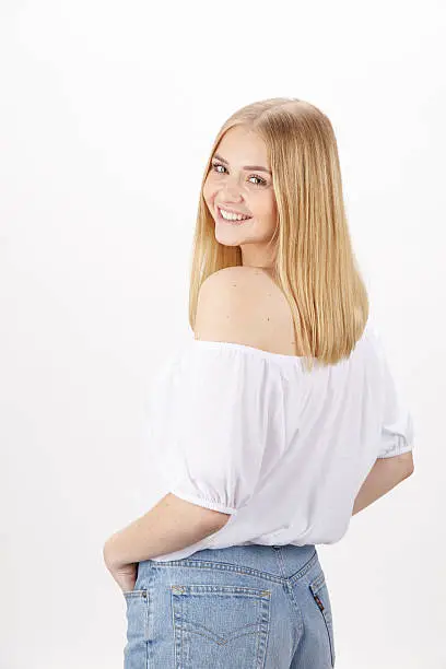 An attractive young blond woman wears summerly clothes (a white blouse and light blue jeans), looks over her shoulder and enjoys the summer. Studio shot over white background.