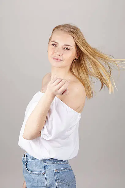 An attractive young blond woman wears summerly clothes (a white blouse and light blue jeans), looks over her shoulder and enjoys a fresh summer breeze. Studio shot over light gray background.