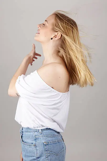 An attractive young blond woman wears summerly clothes (a white blouse and light blue jeans) and enjoys a fresh summer breeze. Studio shot over light gray background.