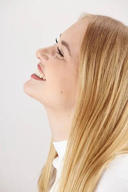 An attractive young blond woman laughs happy. The studio shot shows her face side-face over white background.