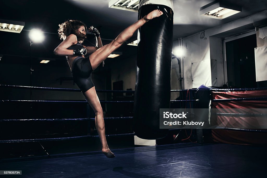 Female kickboxer training with a punching bag Photo of a fit female kickboxer training with a punching bag in a boxing hall. She is jumping on a boxing ring in an underground fight club, training kicking. Kickboxing Stock Photo