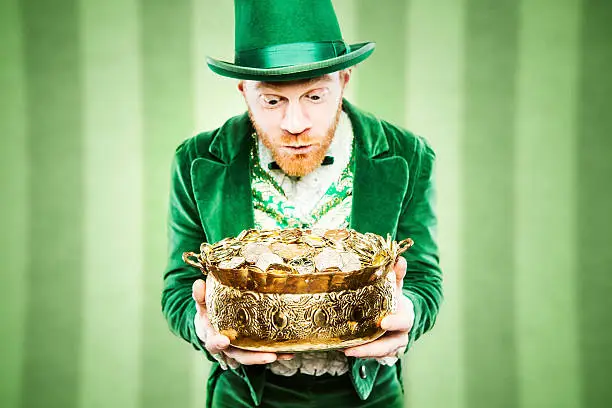 A stereotypical Irish character all ready for Saint Patricks day.  He holds up a pot of gold looking at it with wide excited eyes.  Geen striped wallpaper wall behind him.  Horizontal image with copy space.