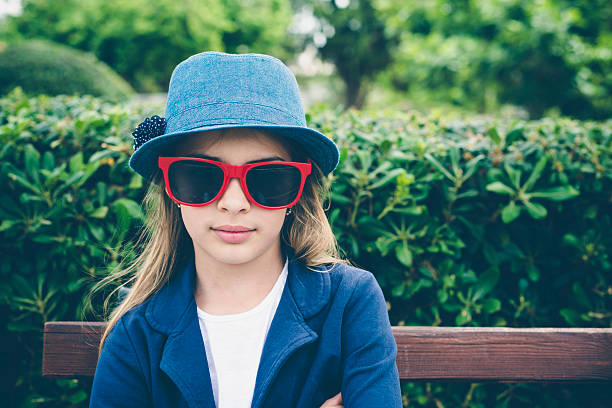 Portrait of the cute little girl in the park with a blue hat and red sunglasses sitting on the bench. Horizontal framing.