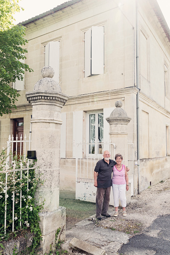 Elderly couple standing in front of their house in summer. Man is tenderly holding his wife by the shoulders. Both are looking at camera with a smile. They are casual dressed in summer clothes. Vertical full length shot with copy space. This was taken in France.
