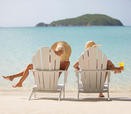 two women in bikinis sitting in white adirondack chairs at a Caribbean beach, enjoying the view and tropical drinks