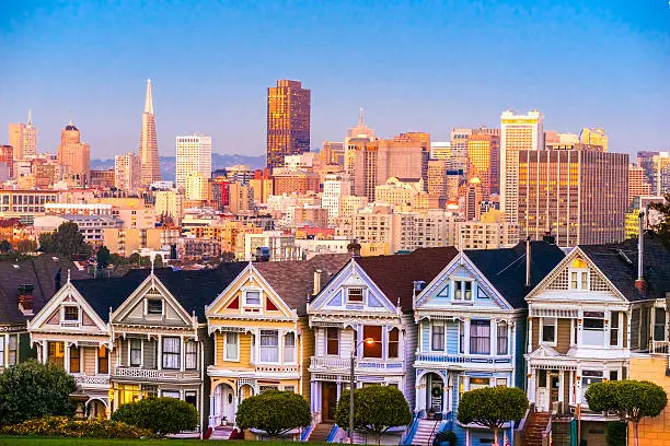 The Painted Ladies of San Francisco, California sit glowing amid the backdrop of a sunset and skyscrapers.