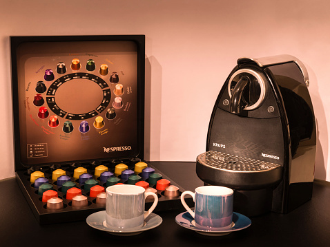 London, England - February 12, 2015: Nespresso is set to open its first cafes in prime locations across Europe, The Nespresso brand endorses machines that brew espresso from its coffee capsules. This studio image depicts a Krups / Nespresso coffee machine and Nespresso boxed capsule coffee set in the background, with two coffee cups and saucers in the foreground. Lighting is focused on the Krups / Nespresso coffee machine.