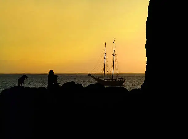 A woman with a dog enjoys the sunset at the sea against the backdrop of a sailboat.