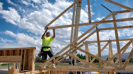 Manual laborer poised on a new construction positioning a piece of frame work against a partly cloudy blue sky.