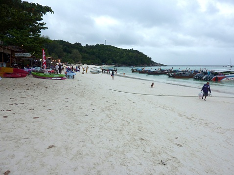 Koh Lipe, Thailand - November 22, 2013: Rainy day on Pattaya beach, Koh Lipe island, a developed long stretch of white sand beach on the Andaman Sea. Local man carrying two sacks walking on the shore. in the background leaving people waiting to be embarked on speed boats
