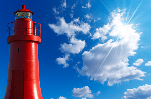 Red metallic lighthouse in the La Spezia harbor, Liguria, Italy. On blue sky with clouds and sun rays