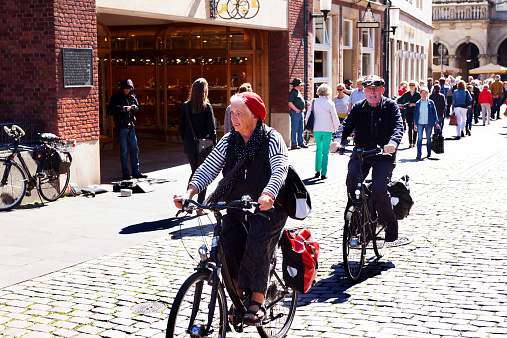 Münster, Germany - May 31, 2014: Capture of a cycling caucasian senior couple in Münster. Couple is coming from street Prinzipalmarkt and is on way to square arounf cathedral. In background people are walking. In front of building is a beggar and homeless man.