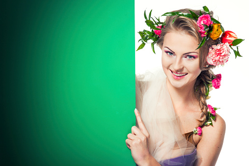 Portrait of a beautiful young blonde with flowers woven into her hair, smiling from behind a green background.