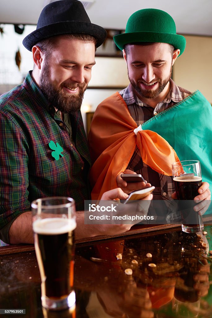 Sending sms greetings Two men text messaging on their smartphones while celebrating St Patricks Day at bar Portable Information Device Stock Photo