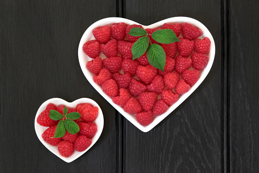 Raspberry fruit in heart shaped bowls with leaf sprigs over wooden black background.
