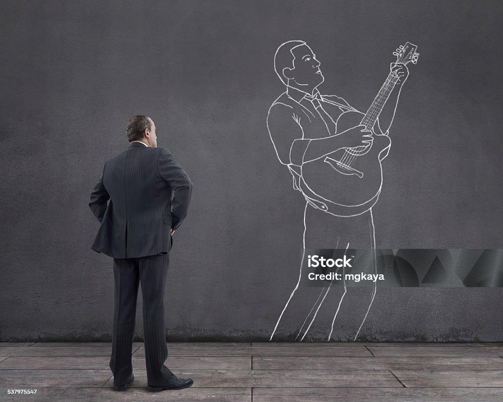 Businessman and Guitarman Rear view of businessman standing on wooden floor with businessman playing acoustic guitar sketched (chalk drawing) on the wall. Acoustic Guitar Stock Photo