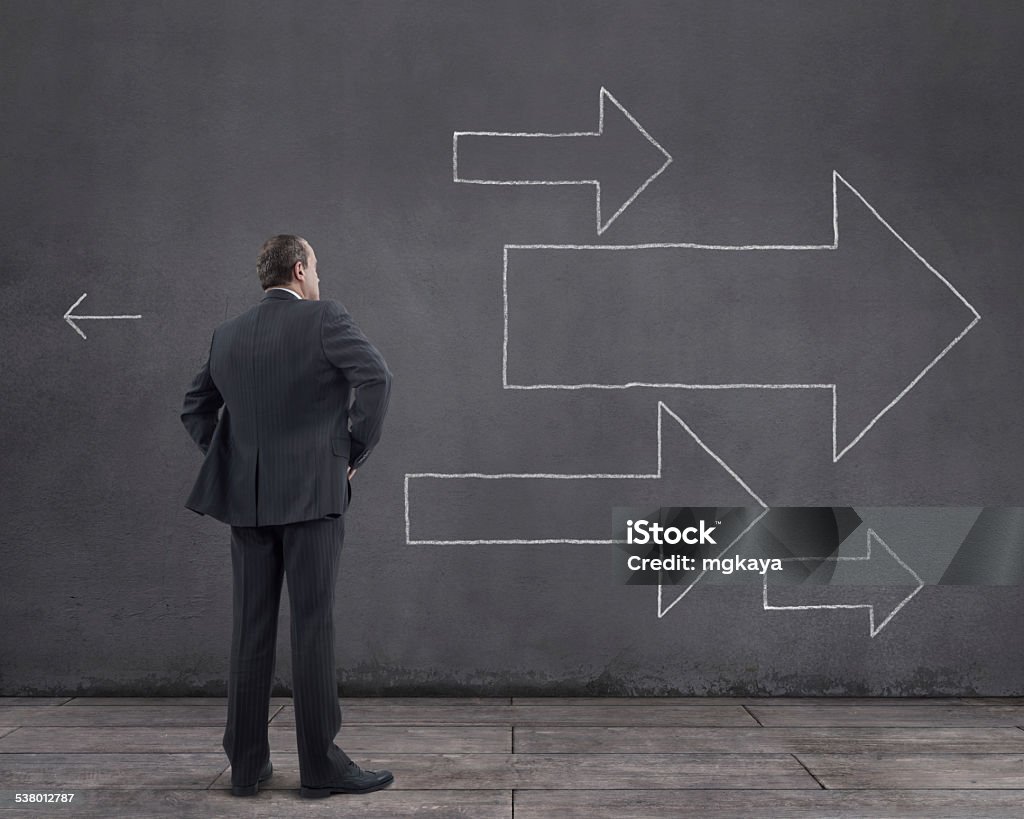Businessman and Arrow Signs Rear view of businessman standing on wooden floor with arrow signs sketched (chalk drawing) on the wall. He is looking further, thinking, searching new ideas and way to exit. 2015 Stock Photo