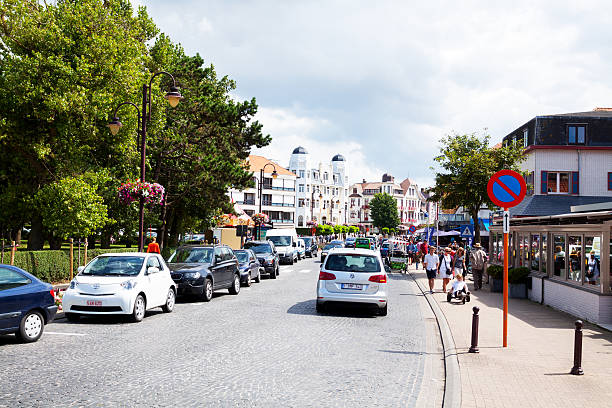 De Haan, Belgium - July 29, 2012: View along street in De Haan in summer. Cars are parked in street. Some traffic is ruling. People are walking along sidewalks. In background are buildings. At ;eft side are trees.