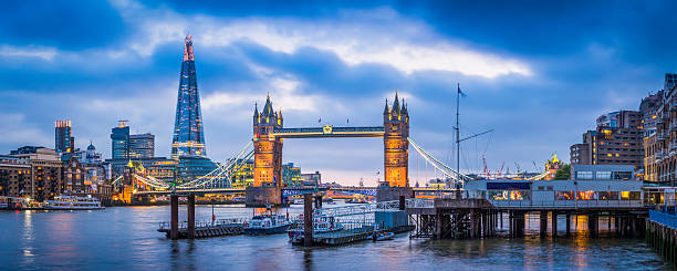 London Tower Bridge and The Shard illuminated over Thames panorama Panoramic view across the River Thames to the historic span of Tower Bridge illuminated against the blue dusk sky overlooked by the futuristic glass spire of The Shard in the heart of London, Britain's vibrant capital city. ProPhoto RGB profile for maximum color fidelity and gamut. gla building stock pictures, royalty-free photos & images