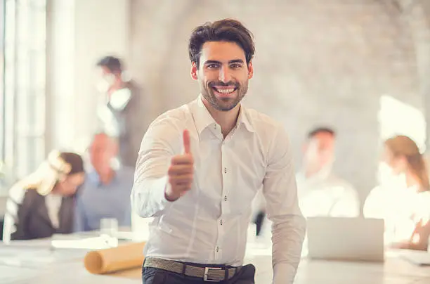 Happy businessman showing thumbs up while sitting in the office and looking at the camera. There are people in the background.