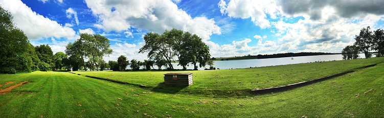 Panorama of the lakeside. This image was taken during the summer. It shows a sunny day. The colours of this image are bright, bold and colourful. This adds colour to the picture. The sun is also reflected off the clouds, this adds colour to the sky and helps to make the sky look blue. 