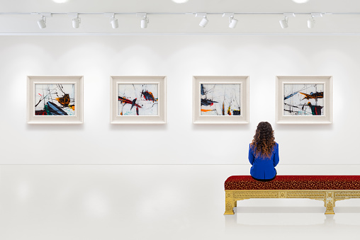 In a exhibition centre, lonely young woman visits an art exhibition and watches artist's collection on the wall. Lightened white wall contains four white frames with artist's painting.